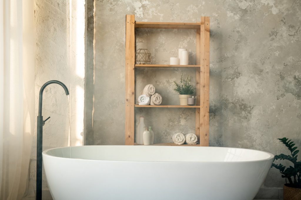 Large porcelain white bathtub and wooden shelves with rolled towels, plastic jars and candles against grey wall in bathroom